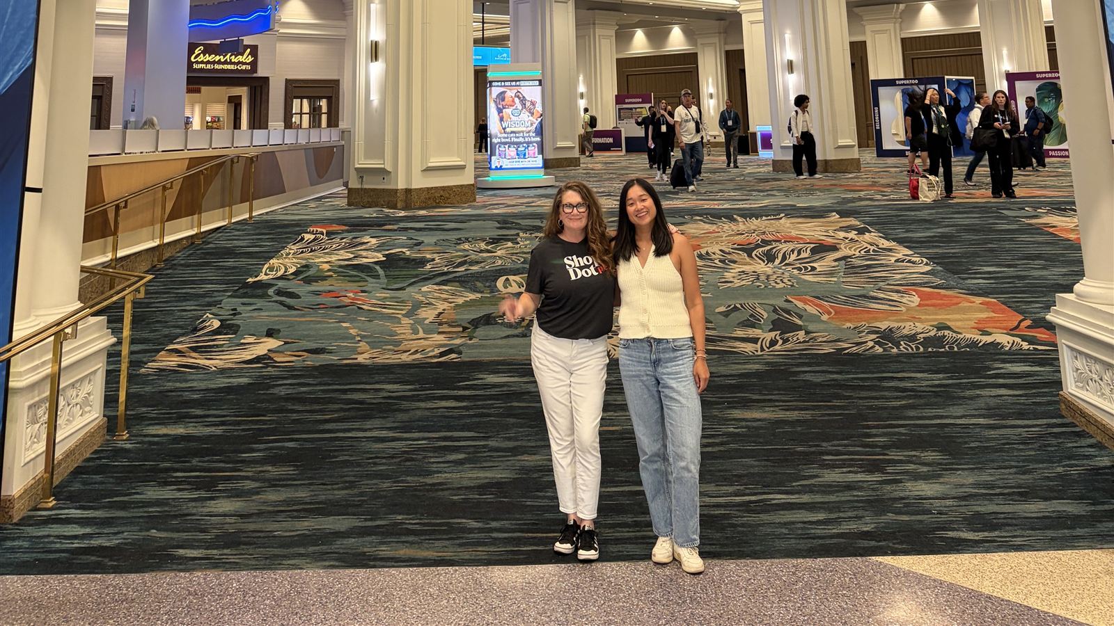 Two ShopDot Pet team members on the SuperZoo 2026 show floor, with booths and attendees in the background.